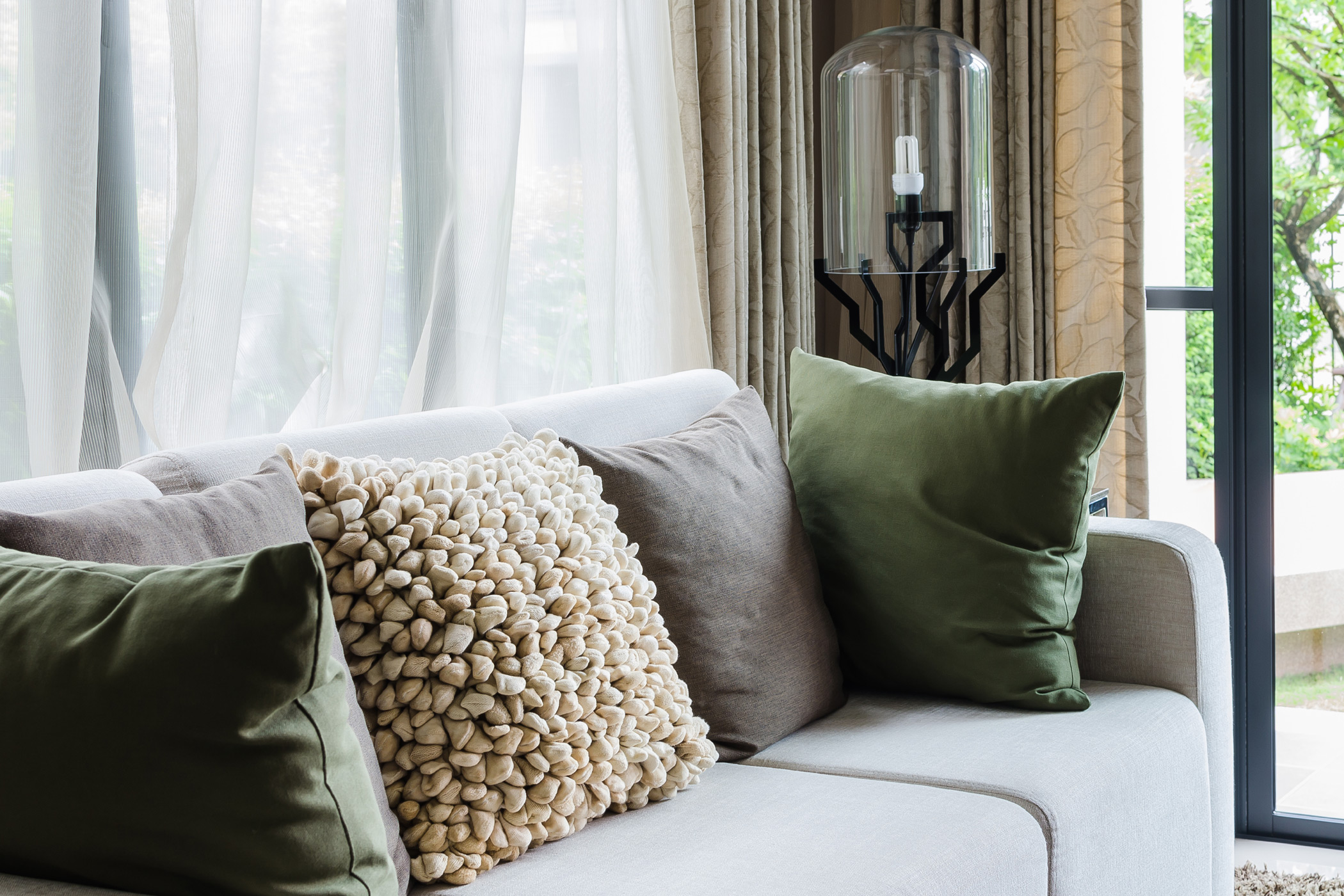a couch covered in throw pillows in front of a window with soft and breezy curtains